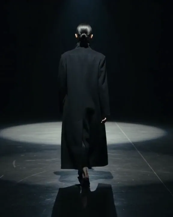 A model in a long black coat walks away under a single spotlight on a dark polished floor.