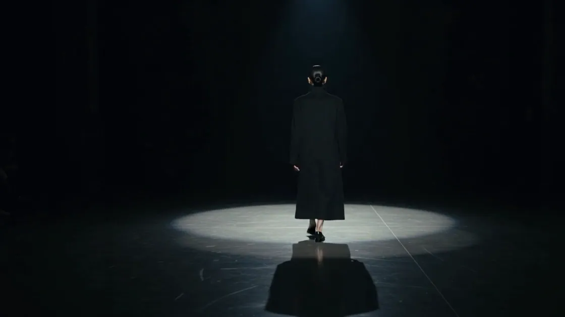 The same model, far off under the spotlight, reflected in the black floor.