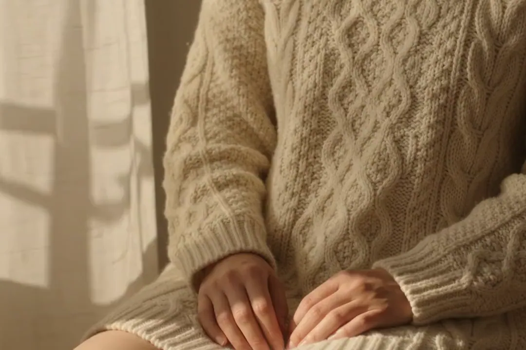 Hands resting in the lap of a model wearing a cream cable-knit sweater.
