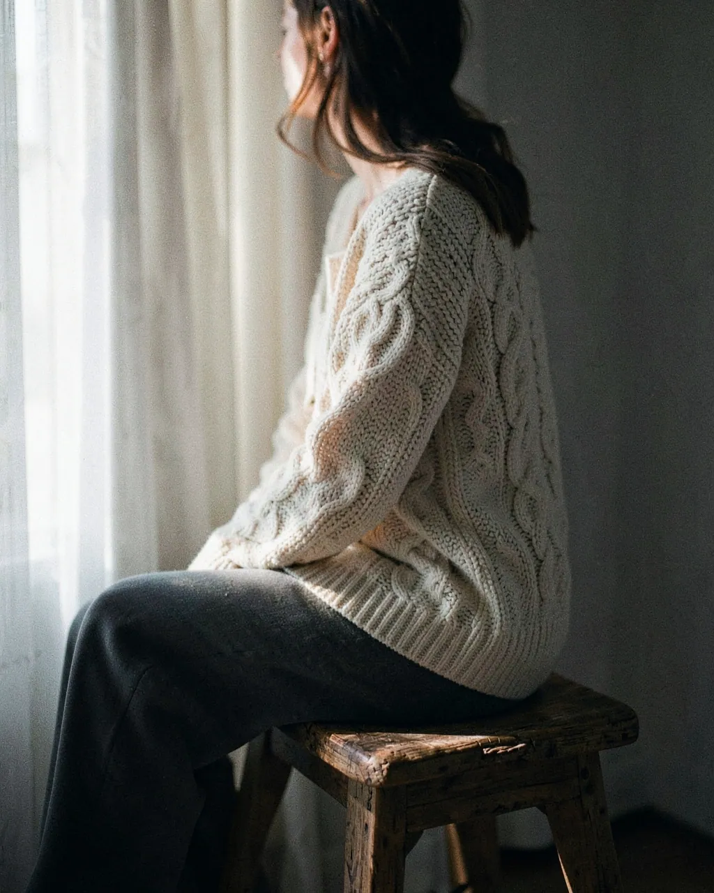 A model in a cream cable-knit cardigan sits by a tall window.