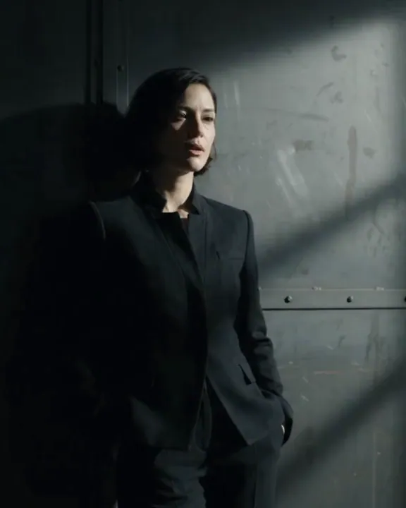 A model in black tailoring leans against a riveted steel door in slatted light.