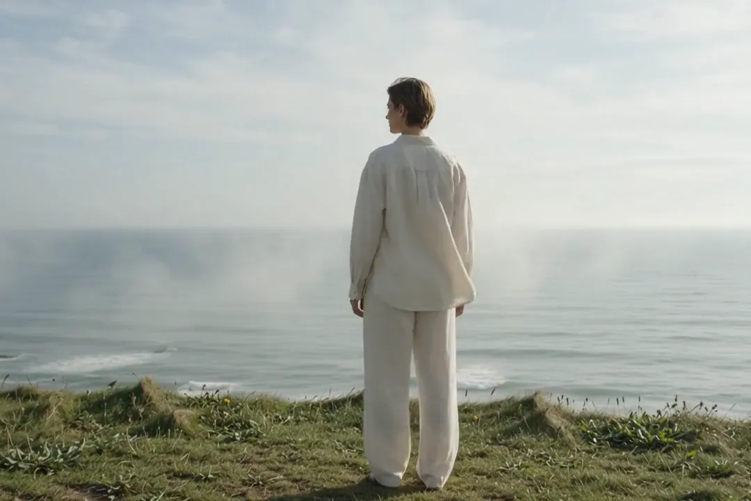A model in cream linen stands on a grassy clifftop looking out at a misty sea.
