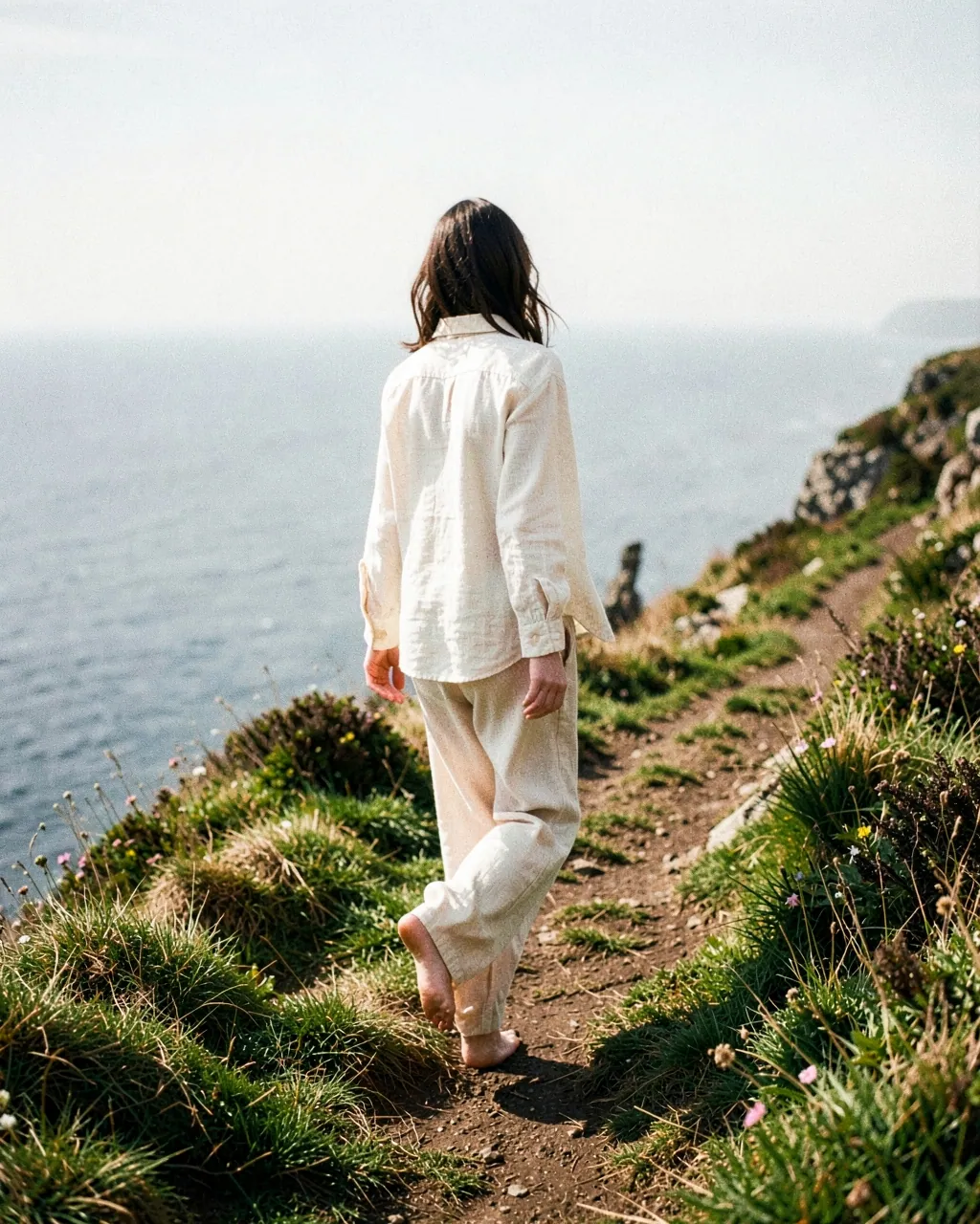 A model in cream linen walks barefoot along a clifftop path above the sea.